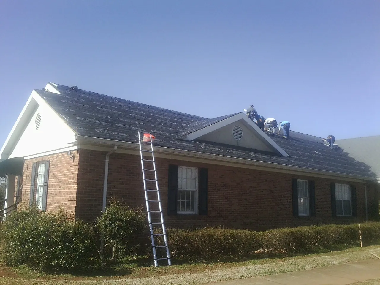 Crew tearing shingles off a brick ranch roof with a ladder set — Emergency Roof Repair work by our crew in East Norwich