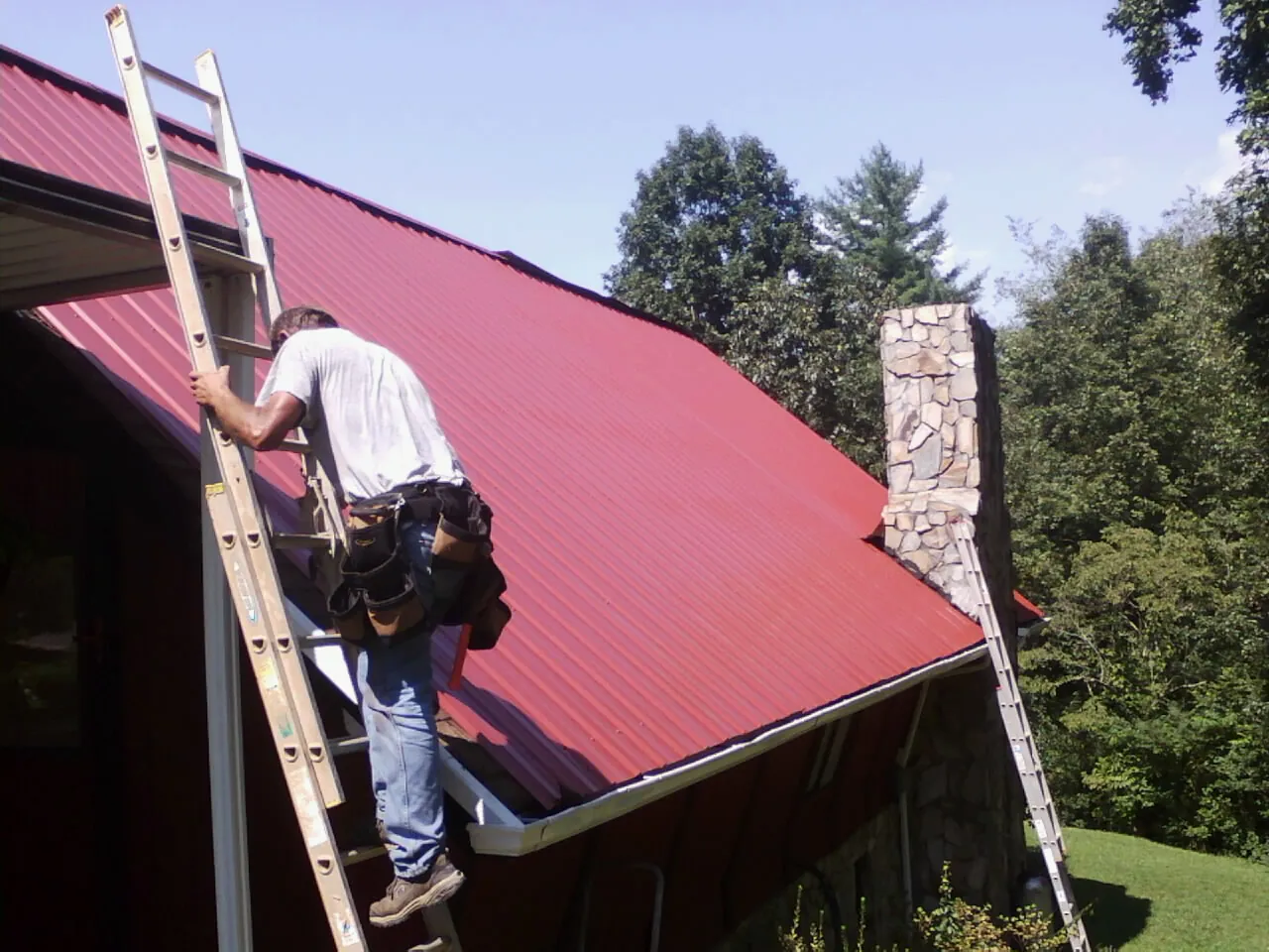Roofer on a ladder beside a red metal roof and stone chimney — Soffit & Fascia Repair by Roofing Brothers in East Norwich